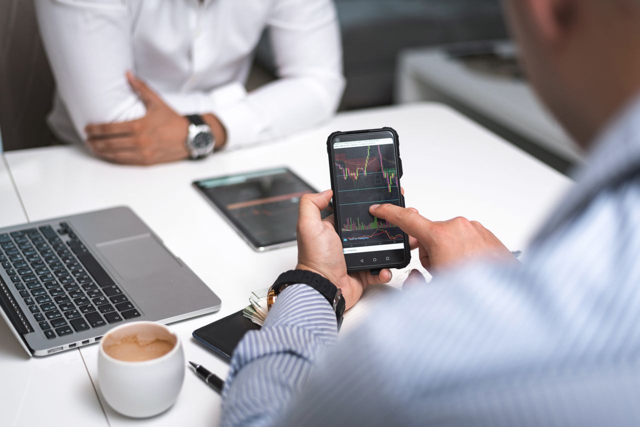 gallery-03 Two businessmen analyze stock market data using a smartphone and laptop in a modern office setting.